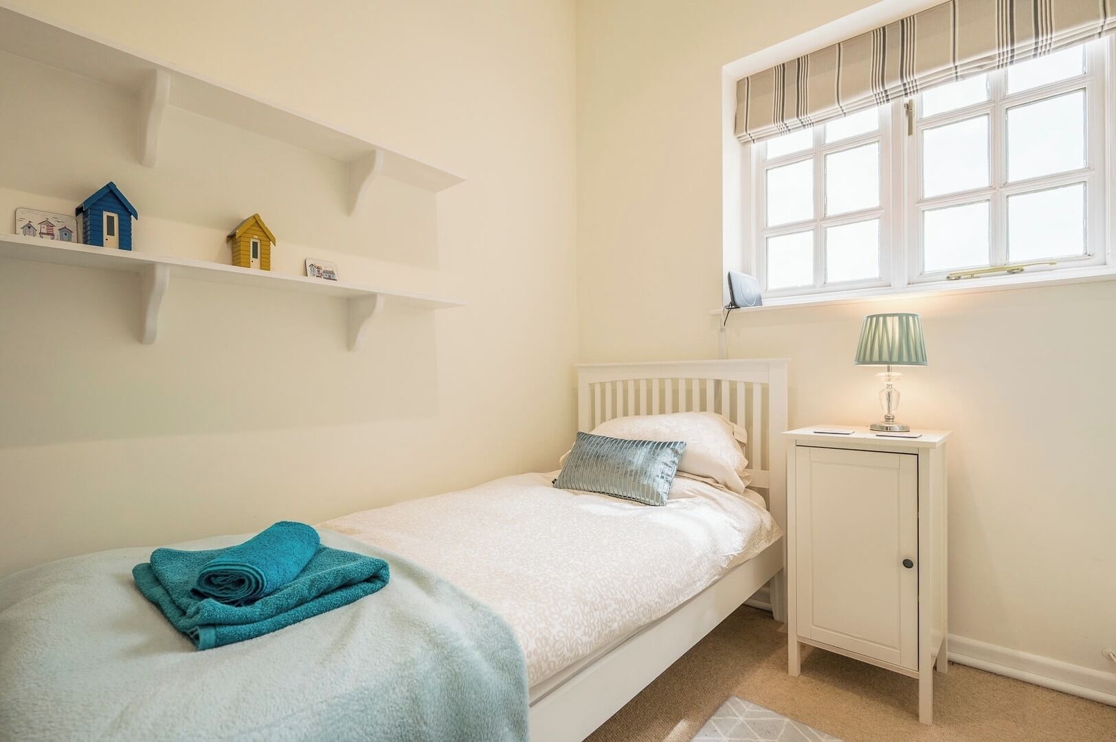 A bedroom with a single bed, a nightstand, a window with blinds, and wall-mounted shelves with decorative items.