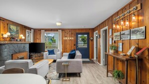Een gezellige woonkamer met een open haard, grijze banken, een houten wand met hangende lampen, en een deur met een blauwe omlijsting.