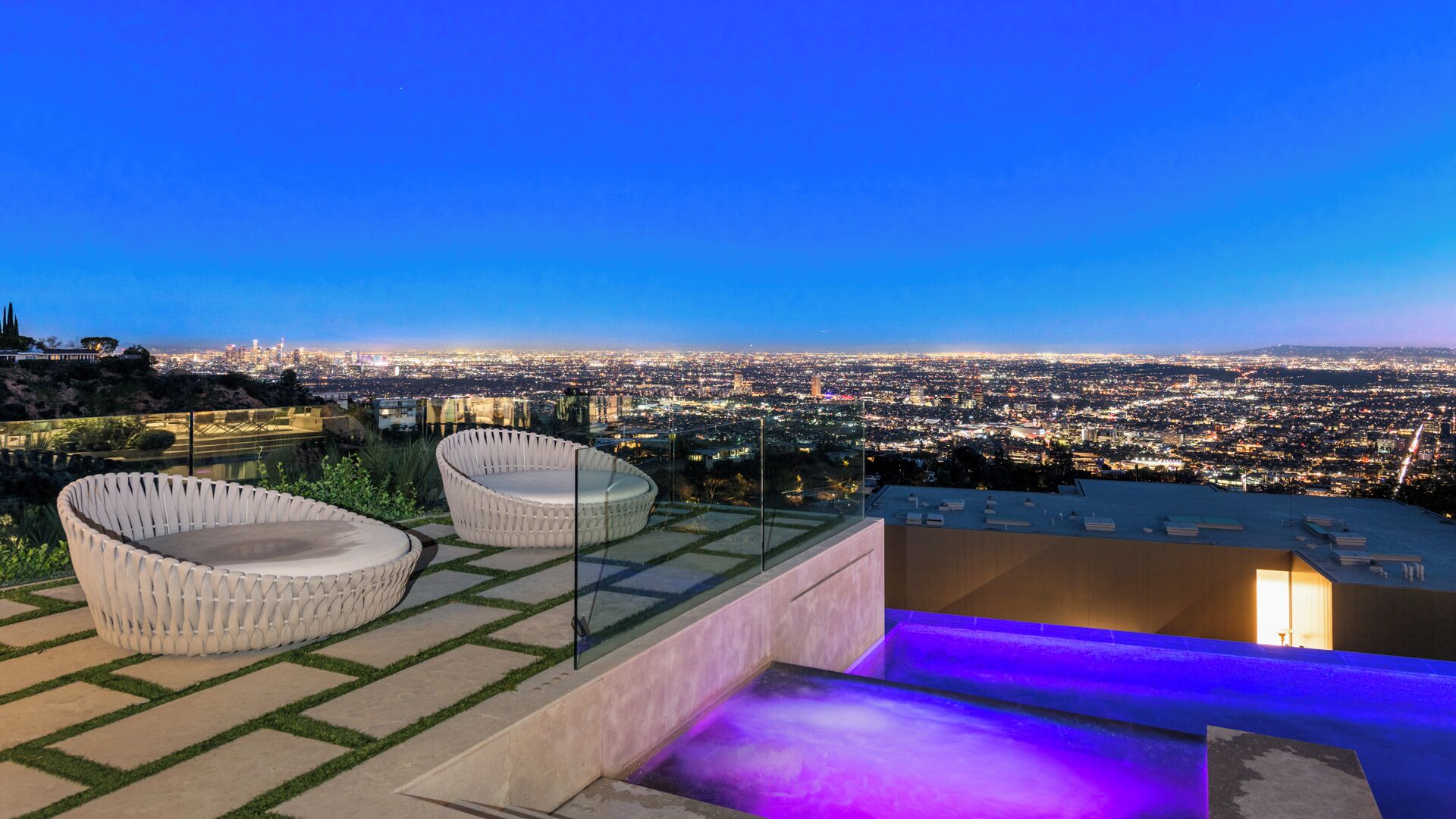 Eine Dachterrasse mit zwei weißen Sitzgelegenheiten, einem kleinen Pool mit violetter Beleuchtung und Blick auf die Stadt bei Einbruch der Dunkelheit.