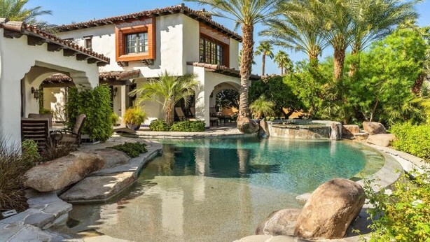 A Mediterranean-style villa with a swimming pool, palm trees, and a stone pathway. - Lakefront 6BR Estate | Private Beach, Pool & Dock (Indio)