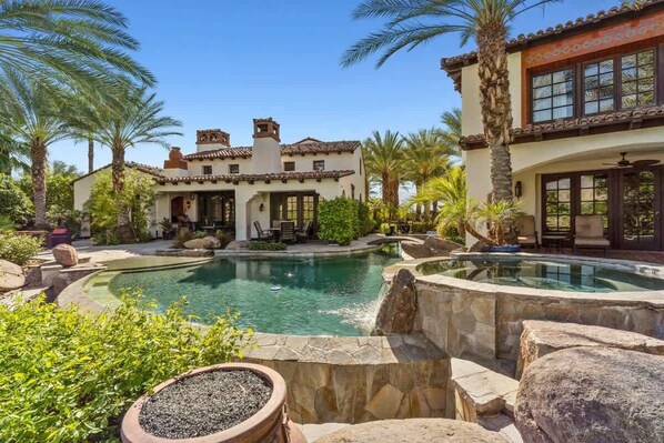 A pool with a hot tub, surrounded by palm trees and a house with a tiled roof. - Lakefront home w/ beach + dock (Indio)
