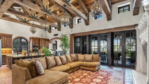 A spacious living area with a large sectional sofa, a patterned red rug, and a high ceiling with exposed beams and hanging lights. - Lakefront 6BR Estate | Private Beach, Pool & Dock (Indio)