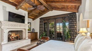 A bedroom with a fireplace, a large bed, a wooden ceiling, and a view through a glass door. - Lakefront 6BR Estate | Private Beach, Pool & Dock (Indio)