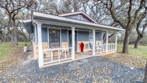 A covered porch with stone walls and a red roof, surrounded by trees. - Live Oak Cottage in Concan (Concan)