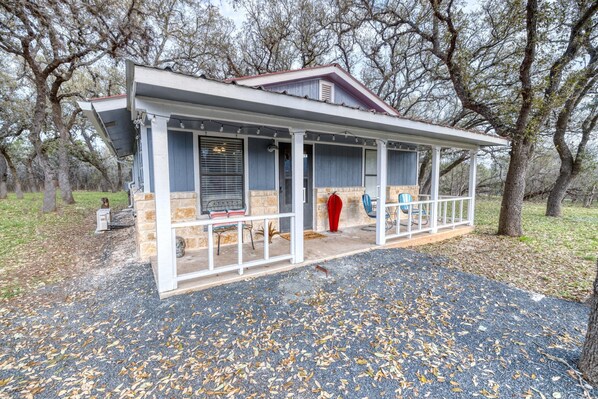 A covered porch with stone walls and a red roof, surrounded by trees. - Live Oak Cottage in Concan (Concan)
