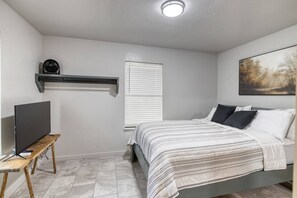 A bedroom with a bed, a TV on a stand, a window with blinds, and a painting on the wall. - Live Oak Cottage in Concan (Concan)