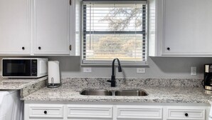 A kitchen with white cabinets, a granite countertop, a stainless steel sink, a microwave, and a window with blinds. - Live Oak Cottage in Concan (Concan)