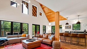 A spacious living area with a large sofa, ottoman, and a red patterned rug. There are multiple windows providing natural light and views of the outdoors.