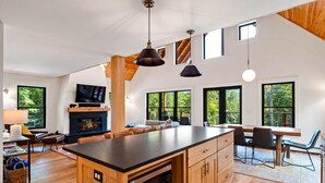 A modern kitchen with a central island, wooden cabinets, and a built-in oven. There are large windows providing natural light and views of the outdoors.