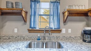 A kitchen with granite countertops, a stainless steel sink, wooden shelves, and a window with blue curtains. - Oasis Cabin #2 on the Frio (Concan)