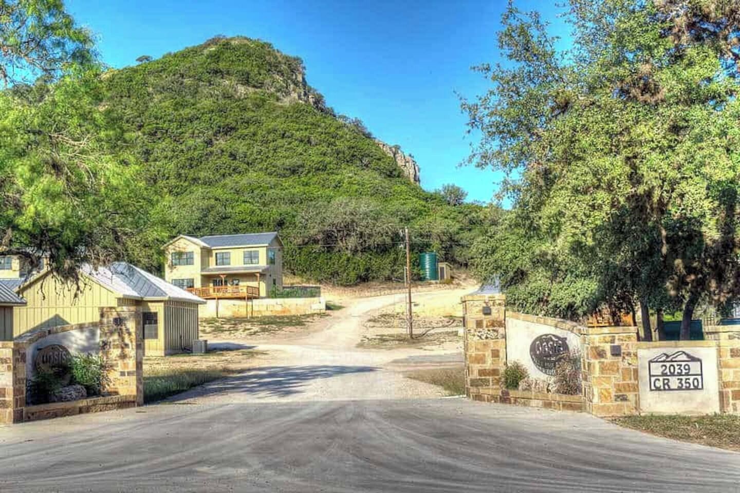 A residential area with a paved road leading to a gated entrance. There are houses with visible driveways and a mountain in the background.