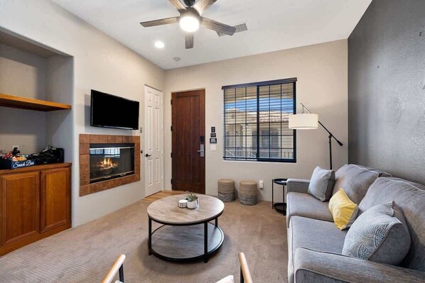A living room with a fireplace, a sofa, a coffee table, a TV, and a ceiling fan. - Old Town La Quinta | Pool-View Terrace + Hot tub (La Quinta)