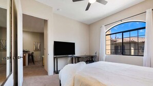 A bedroom with a bed, a desk with a flat-screen TV, a ceiling fan, and a large window with curtains. - Old Town La Quinta | Pool-View Terrace + Hot tub (La Quinta)