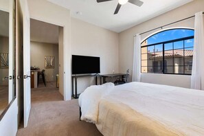 Une chambre à coucher avec un lit, un bureau, une télévision et une grande fenêtre avec des rideaux.