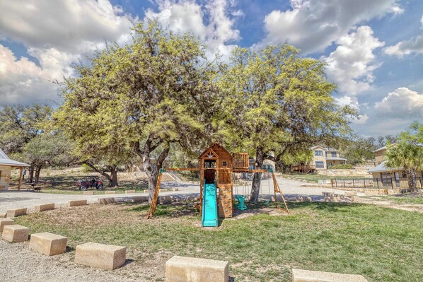 A playground with a wooden playhouse and slide, surrounded by trees and open grassy areas.