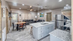 A spacious kitchen with a central island, a dining area with a round table and chairs, and a living area with a sofa and a TV. - River’s Edge Cabin #3 at Oasis Resort (Concan)