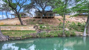 A modern house with a large tree in front, a stone retaining wall, and a small pond. - River’s Edge Cabin #3 at Oasis Resort (Concan)