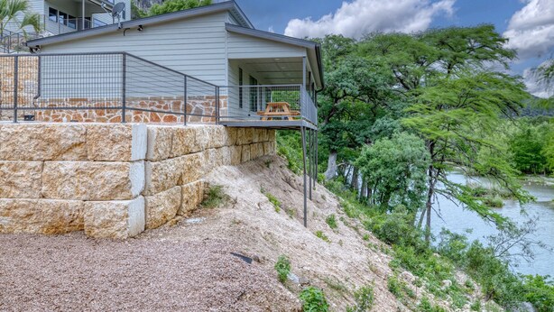A house with a balcony overlooking a river, surrounded by greenery and a stone retaining wall. - Oasis at River’s Edge Cabin #4 (Concan)