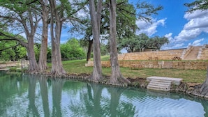 A tranquil outdoor setting with a pond, large trees, picnic tables, and stone steps leading to a viewing area. - River's Edge Lodge #1 on the Frio (Concan)