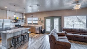 A modern kitchen with a central island, bar stools, and a dining area with a brown leather couch. - River's Edge Lodge #1 on the Frio (Concan)