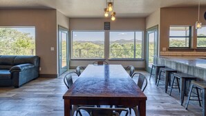 A spacious room with a long wooden table, multiple chairs, and a bar area. Large windows offer a view of the outdoors. - River's Edge Lodge #2 on the Frio (Concan)