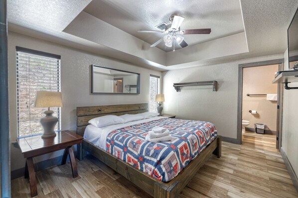 A bedroom with a wooden bed, a ceiling fan, a window with blinds, a bedside table with a lamp, and a bathroom visible through an open door. - River's Edge Lodge #3 on the Frio (Concan)