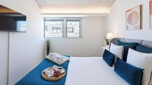 A hotel room with a bed, a bedside table with a lamp, a window with a view of buildings, and a tray with breakfast items on a blue bed runner.