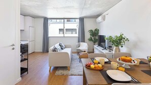 A modern living room with a sofa, dining table, and a TV. There is a kitchen area with cabinets and a refrigerator.