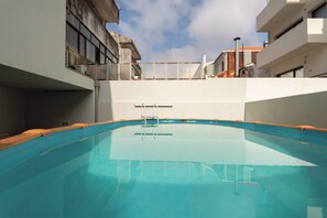 Piscina com deck de madeira, cercada por edifícios com varandas e um céu claro.