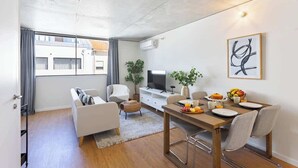 A modern living room with a sofa, dining table, and chairs. There is a TV, a plant, and a painting on the wall.