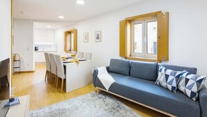 Een moderne woonkamer met een bank, een eettafel en stoelen. Er is een raam met houten kozijnen en een deur die naar een andere kamer leidt.