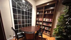 A room with a round wooden table, two chairs, a bookshelf, and a potted plant.