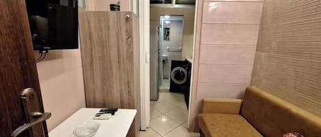 A compact hotel room with a wooden door, a small table with a bowl, a sofa, a washing machine, and a television mounted on the wall.