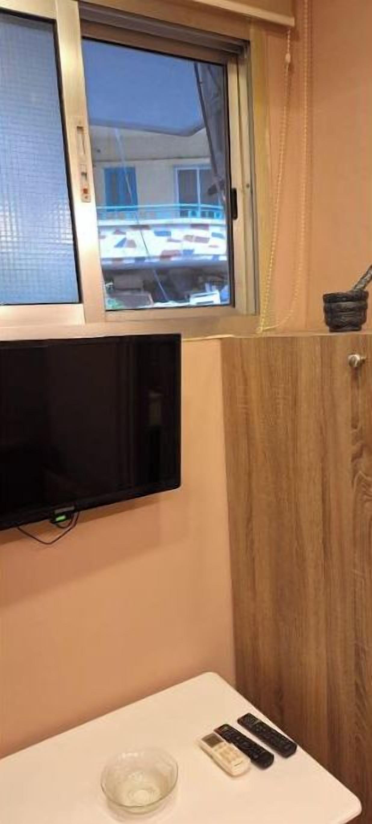 A small room with a television mounted on the wall, a small table with a bowl and remote controls, and a window with a view of a building outside.