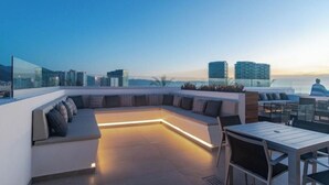 En takterrass med en böjd sittgrupp, ett matbord och utsikt över stadens silhuett i skymningen.