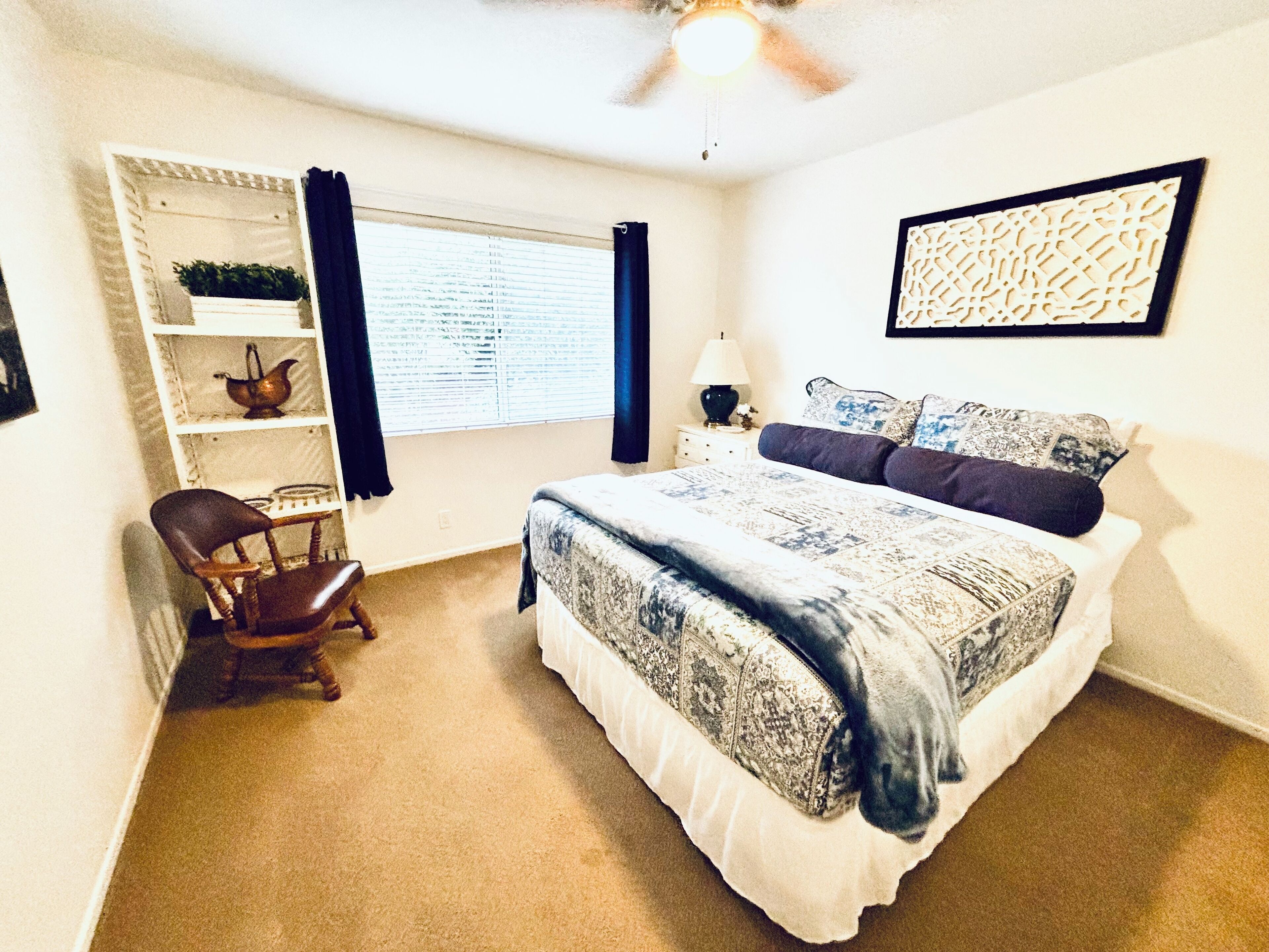 Une chambre à coucher comprenant un lit, une chaise, une fenêtre avec des stores, un ventilateur de plafond et une étagère fixée au mur, ornée d’objets décoratifs.