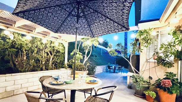 An outdoor patio with a round table and chairs, surrounded by potted plants and a patterned umbrella.