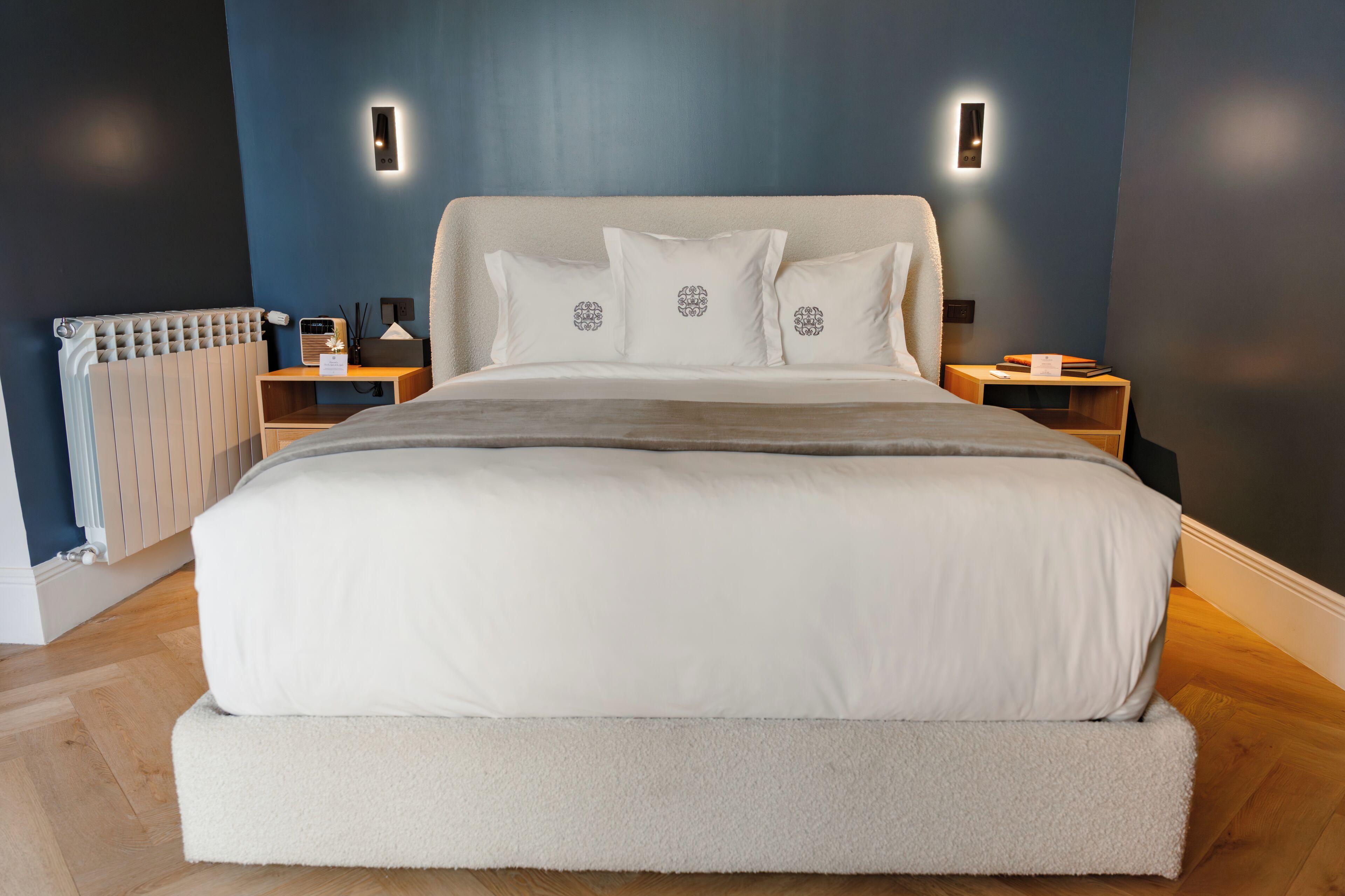 Una cama grande con un colchón blanco mullido y almohadas a juego, flanqueada por dos mesitas de noche con lámparas, en una habitación con paredes azul oscuro y un radiador.
