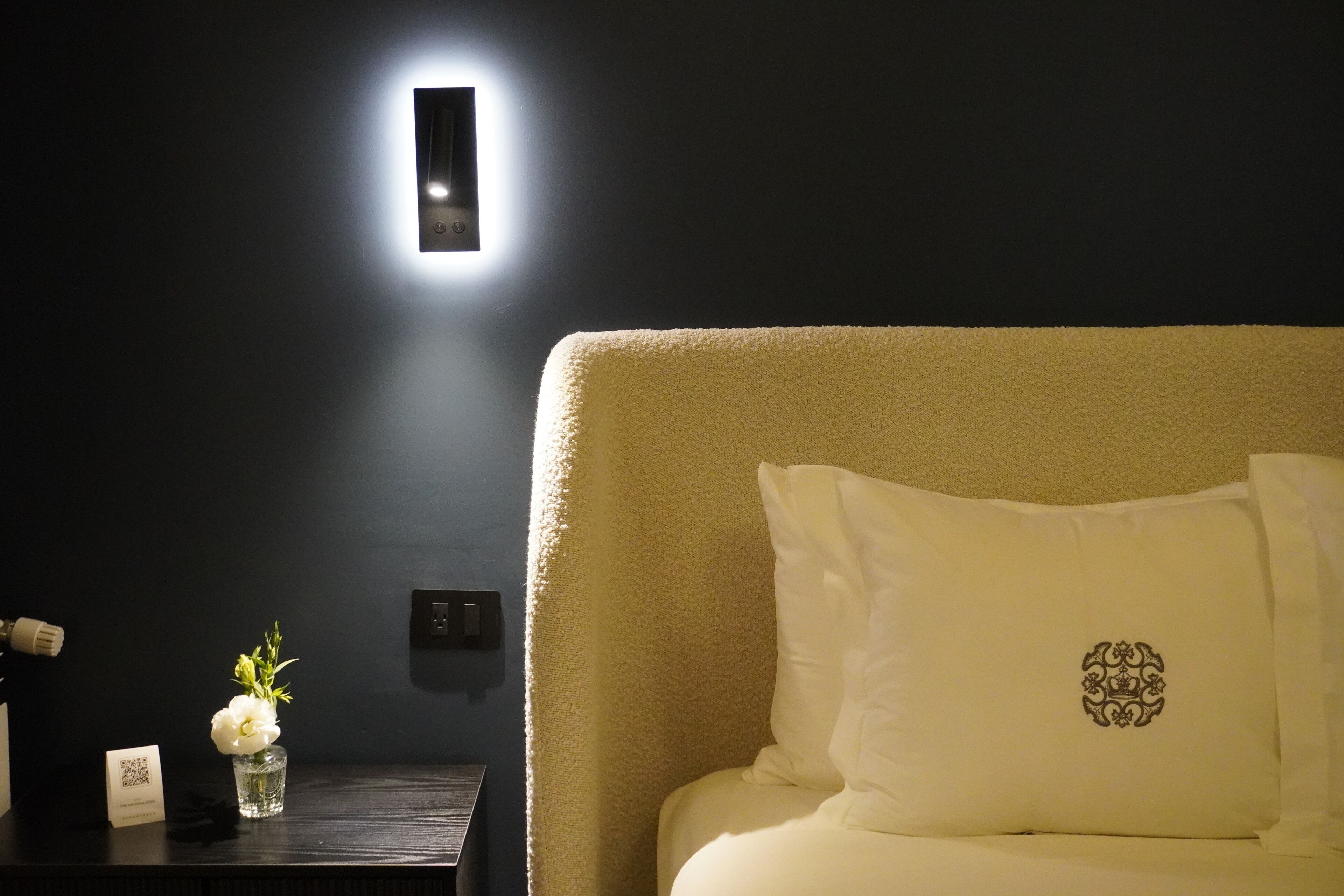 A hotel room with a dark wall, a bedside table with a vase of flowers, a small card, and a bed with a white pillow featuring a crest.