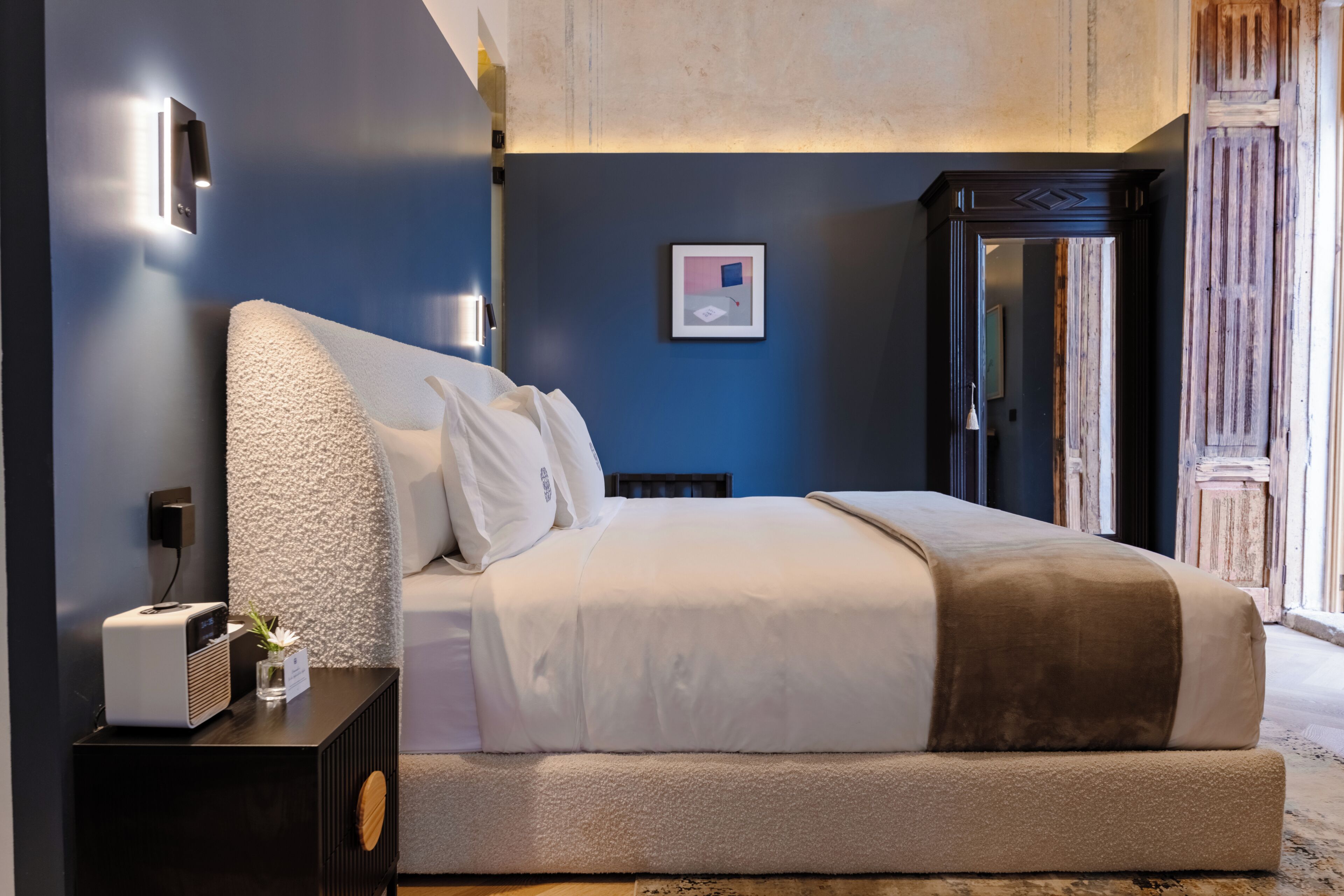 A bedroom with a large bed, a bedside table with a lamp, and a wall-mounted light fixture.