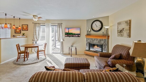 A living room with a fireplace, striped sofas, a dining area with a table and chairs, and a television mounted on the wall. - Coveted DTC Apartment! Free Parking & Fast WiFi! (Greenwood Village)