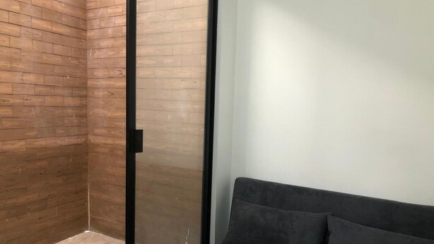 A room with a wooden wall, a glass door, and a grey sofa. - Joint apartments near Bello Ant’s Town Square (Bello)