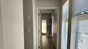 A hallway with a wooden floor, a door at the end, and a window with blinds on the right. - Stylish 2BR Suite in Vibrant LA Area -> Complimentary WiFi, AC & Parking (Los Angeles)