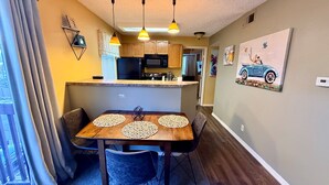 Un coin repas avec une table et des chaises en bois, une cuisine à l’arrière-plan et un tableau accroché au mur.