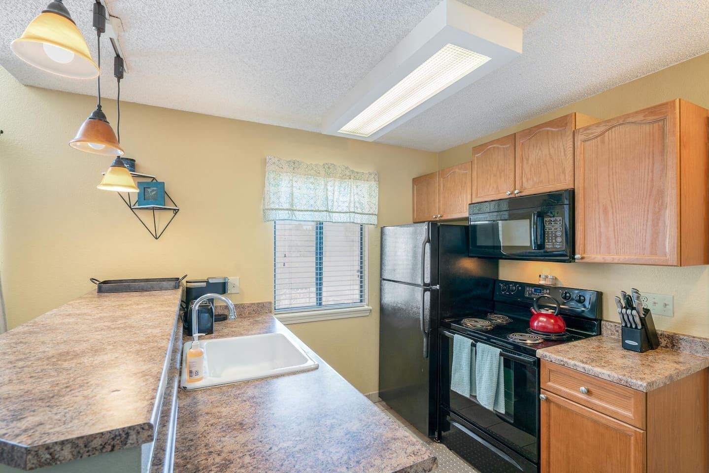 A kitchen with granite countertops, a black refrigerator, a stove, and a microwave. There are wooden cabinets and a window with blinds.