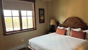 A neatly made bed with white linens, a wooden headboard, and several pillows. A window with blinds allows natural light into the room.