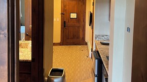 A hallway with a wooden door, a trash can, and a kitchen area with a countertop and sink.