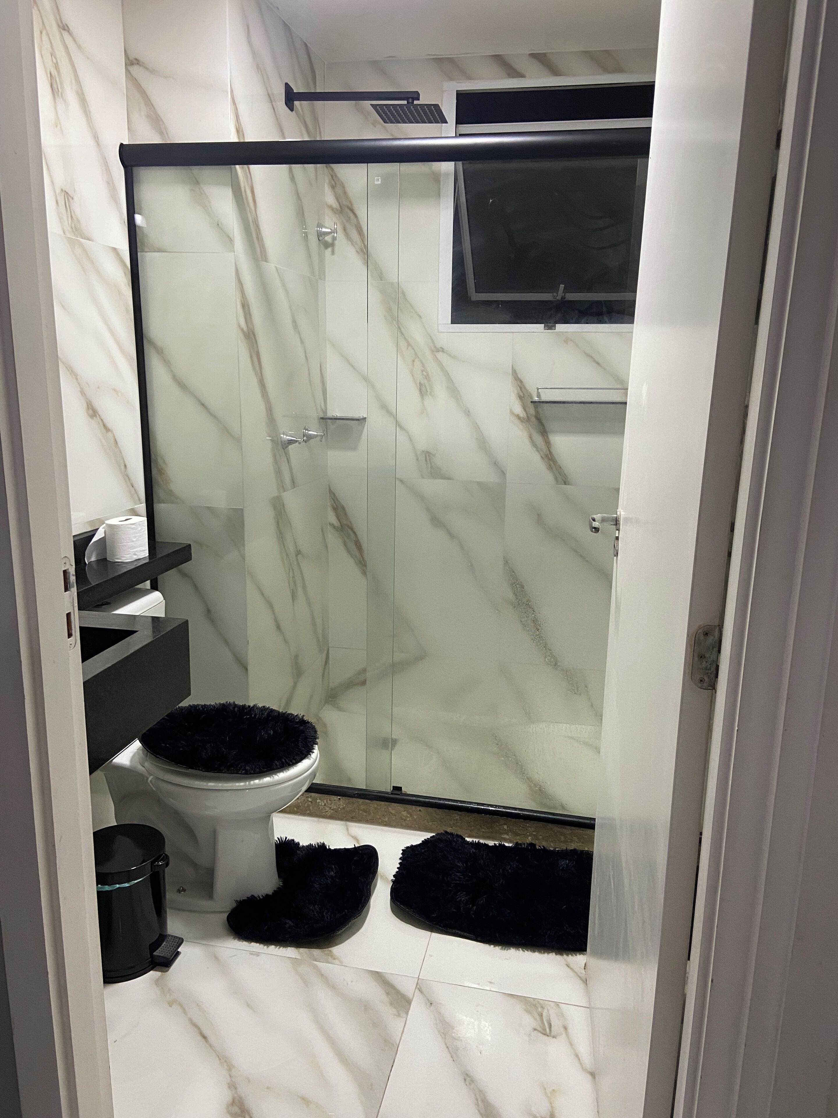 A bathroom with a glass shower enclosure, a toilet, a black trash bin, and a black bath mat.