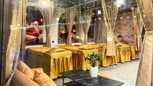 Una habitación con varias camas cubiertas con sábanas doradas, una mesa de mármol negro y luces colgantes tipo guirnalda.
