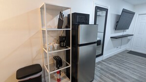 A compact kitchen with a stainless steel refrigerator, a white shelving unit with cups and a coffee maker, and a black countertop microwave. - Fun in the South and Sun (Hallandale Beach)